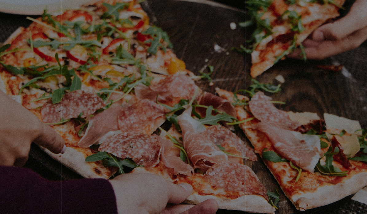A close-up photograph of a pizza topped with prosciutto, salami, arugula and peppers being shared at a wooden table.